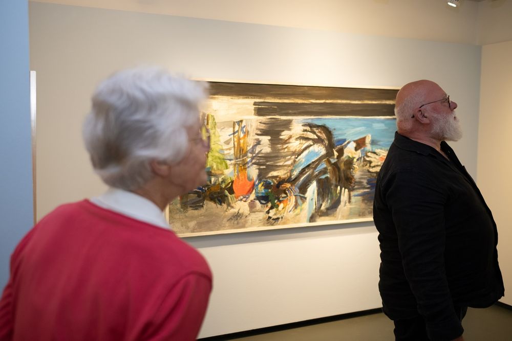 Op de voorgrond staat een oudere vrouw in een rood vest, naast haar een man gekleed in zwart, met op de achtergrond een abstract schilderij.