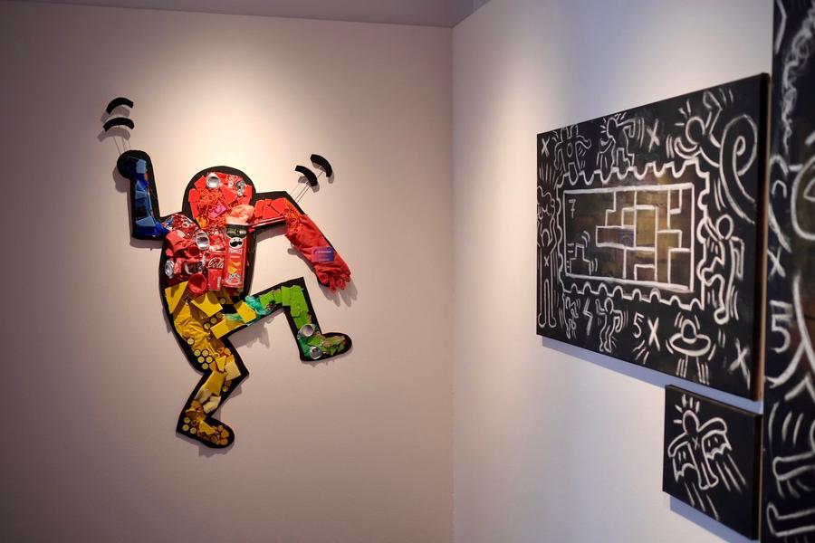 Kunstwerk in de stijl van Keith Haring, opgebouwd uit felgekleurde materialen zoals verpakkingen en blikjes, hangt aan een witte muur naast een zwart schilderij met witte lijnen en figuren.