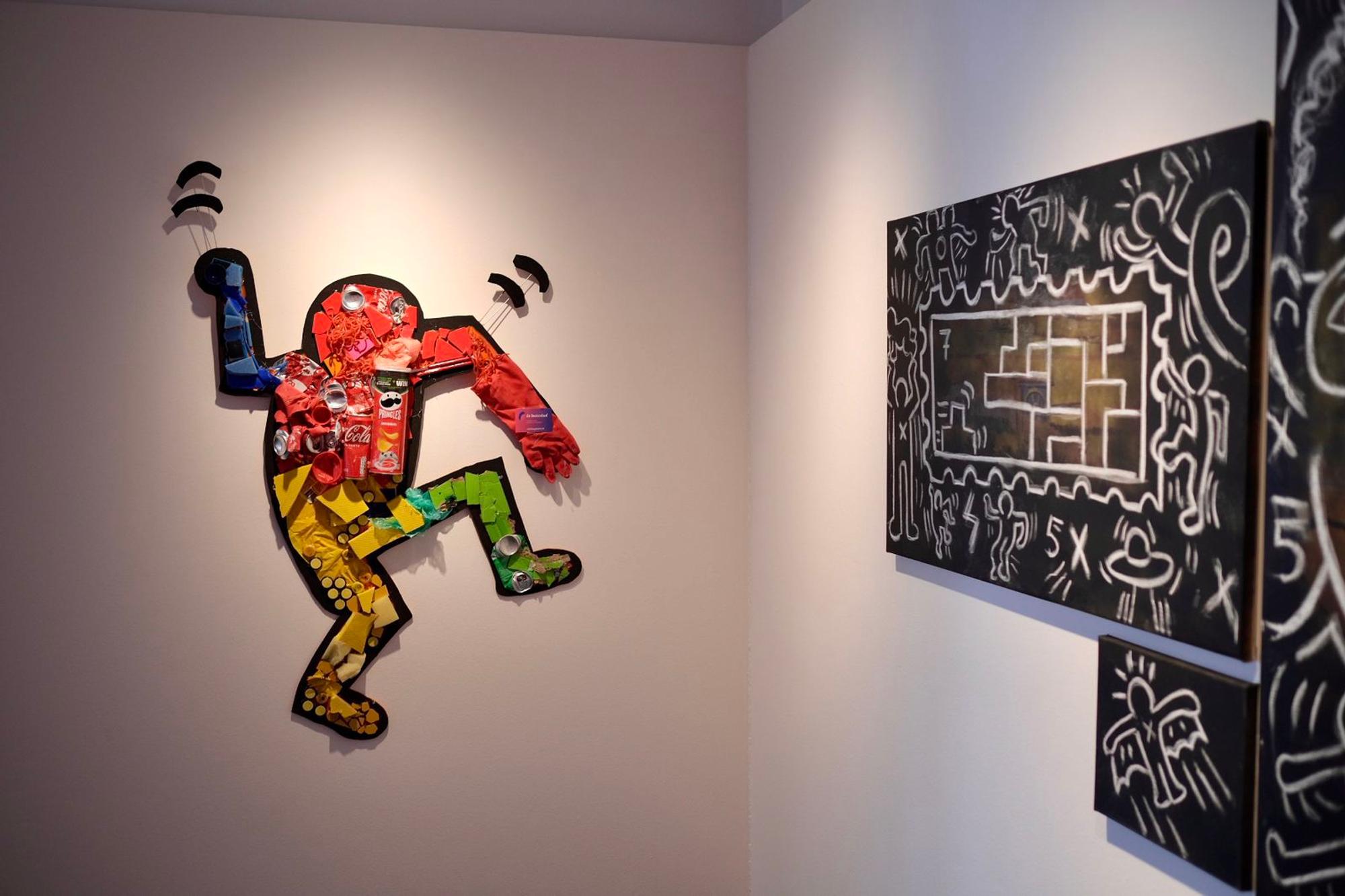 Kunstwerk in de stijl van Keith Haring, opgebouwd uit felgekleurde materialen zoals verpakkingen en blikjes, hangt aan een witte muur naast een zwart schilderij met witte lijnen en figuren.