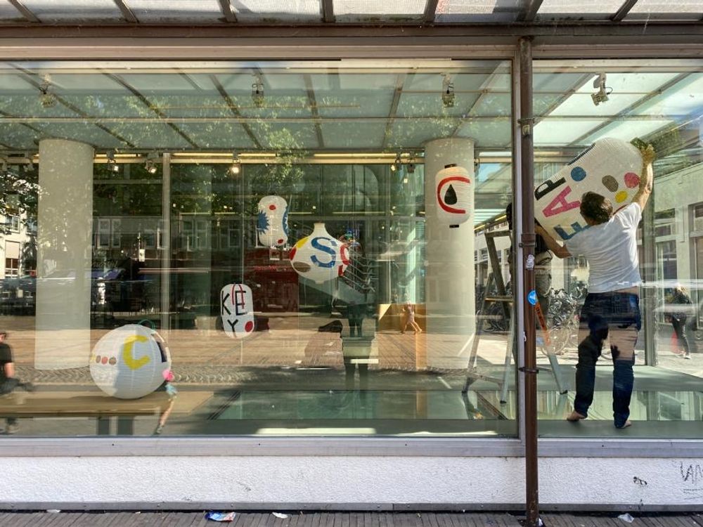 Kunstenaar Jan Rothuizen plaatst handgeschilderde lantaarns in de vitrine van SCHUNCK Glaspaleis in Heerlen, met kleurrijke letters en symbolen zichtbaar op de lampen.