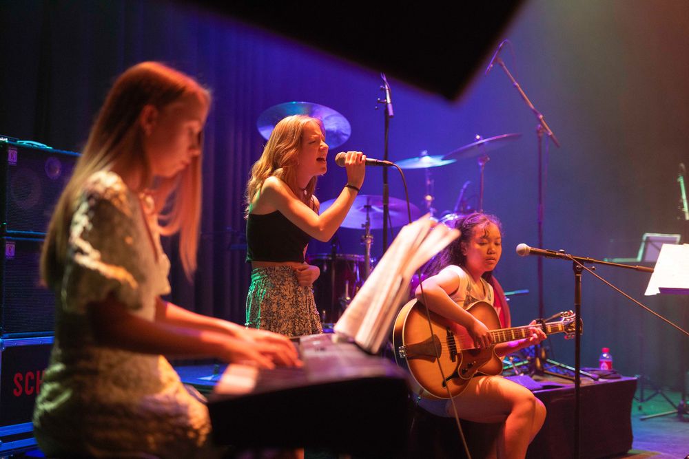 Op een podium tijdens een optreden van SCHUNCK Academy spelen drie jonge muzikanten samen: links wordt een keyboard bespeeld, in het midden wordt gezongen in een microfoon en rechts wordt gitaar gespeeld terwijl er wordt gezongen. Op de achtergrond staan een drumstel en versterkers, en het geheel wordt verlicht met gekleurde podiumlichten in paars, blauw en oranje tinten.
