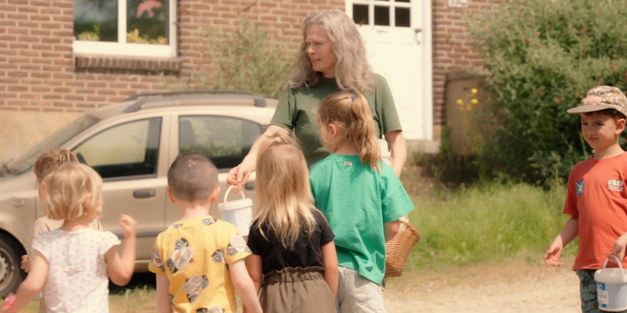 Volwassene staat buiten bij een groep jonge kinderen die mandjes en emmertjes vasthouden, naast een geparkeerde auto bij een stenen gebouw.