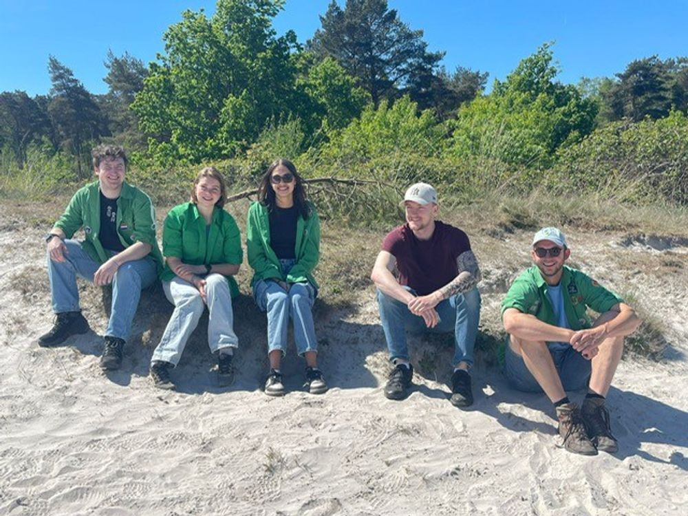 Vijf personen zitten naast elkaar op een zandige ondergrond met groen struikgewas en bomen op de achtergrond.