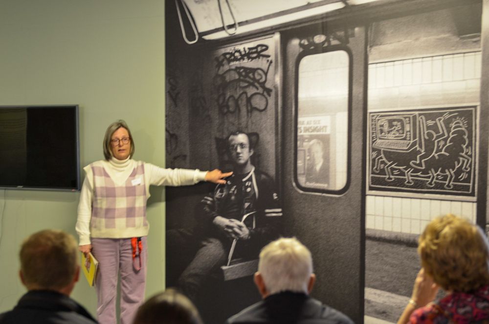 Een persoon die voor een grote zwart-witfoto staat die aan een muur is tentoongesteld. De foto toont een metroscène met graffiti en een advertentieposter. De persoon wijst naar de foto terwijl verschillende mensen zitten en naar de persoon kijken of aandachtig luisteren.