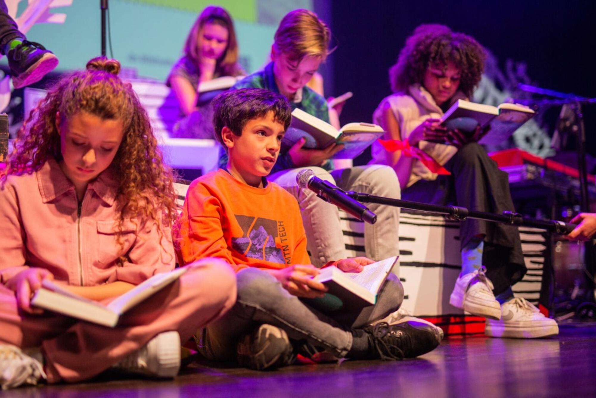 Kinderen zitten op een podium met boeken in de hand tijdens de Nationale Voorleeswedstrijd. Eén jongen in een oranje trui leest hardop voor in een microfoon, terwijl de rest geconcentreerd meeleest in een kleurrijke, theatrale setting.
