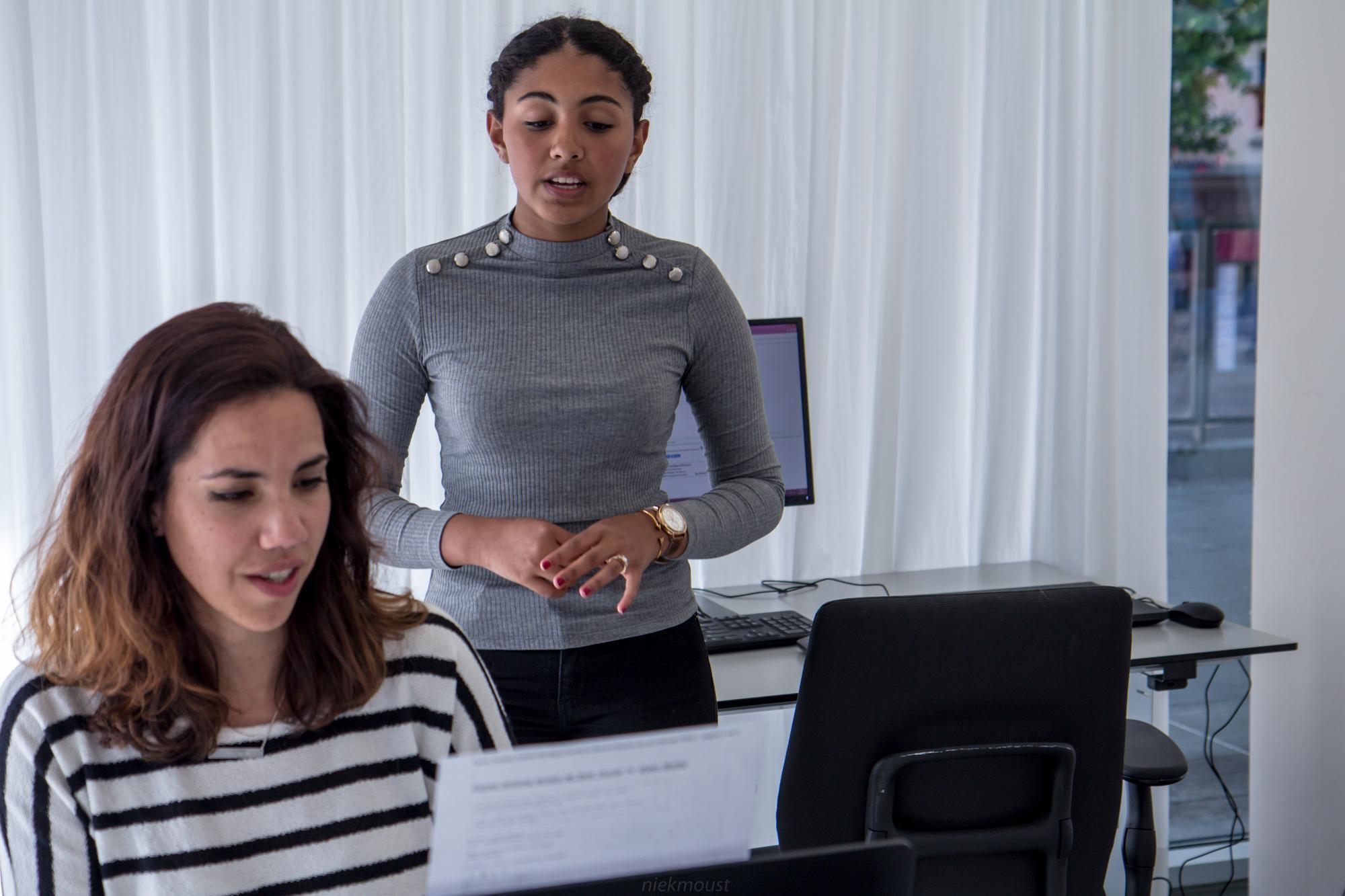 Twee vrouwen in een kantoorruimte; de zittende vrouw leest een document met een geconcentreerde blik, terwijl de staande vrouw iets uitlegt of bespreekt. Op de achtergrond zijn computers en witte gordijnen zichtbaar.