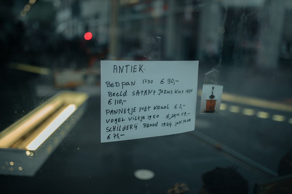 Een handgeschreven kaartje met de tekst "ANTIEK" hangt aan de binnenkant van een vitrine en toont een lijst met objecten, jaartallen en prijzen, zoals een bedpan uit 1730 en een schilderij van Jan Plom uit 1944. Op de achtergrond zijn vage reflecties van het straatbeeld zichtbaar.