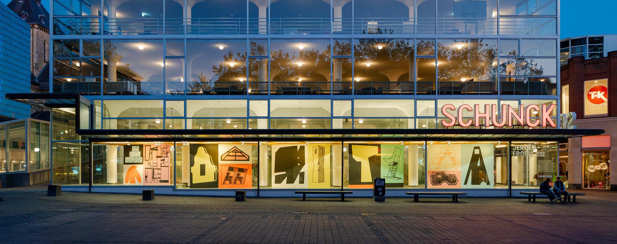 Buitenaanzicht van het Glaspaleis van SCHUNCK in de avond, met verlichte vitrines gevuld met grafische kunstwerken van Jeroen Erosie.