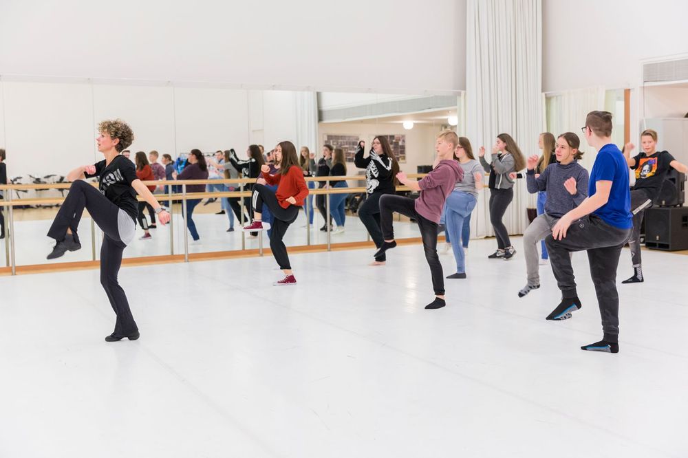 Jongeren volgen een dansles in een lichte danszaal met spiegels; een dansdocent leidt de groep terwijl iedereen met één been opgetild een beweging nadoet.
