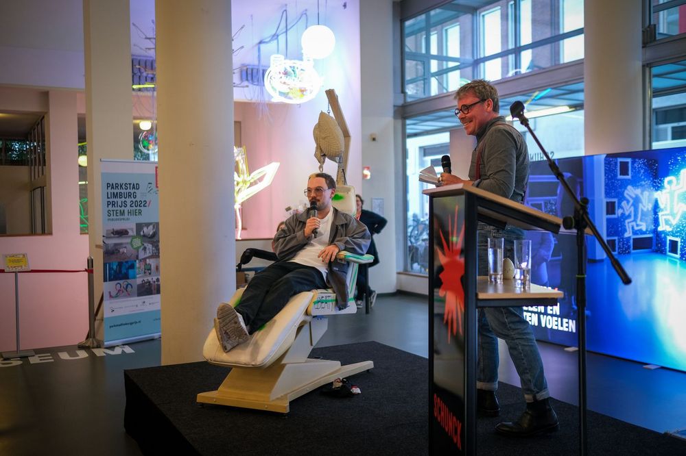 Twee mannen tijdens een presentatie bij SCHUNCK; één zit in een ligstoel met een microfoon en de ander staat lachend achter een spreekgestoelte met een microfoon in de hand.