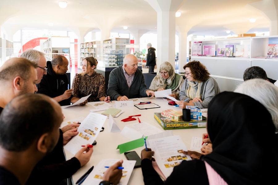 Een groep mensen die rond een tafel zitten. Ze houden een pen en papier vast en helpen elkaar.