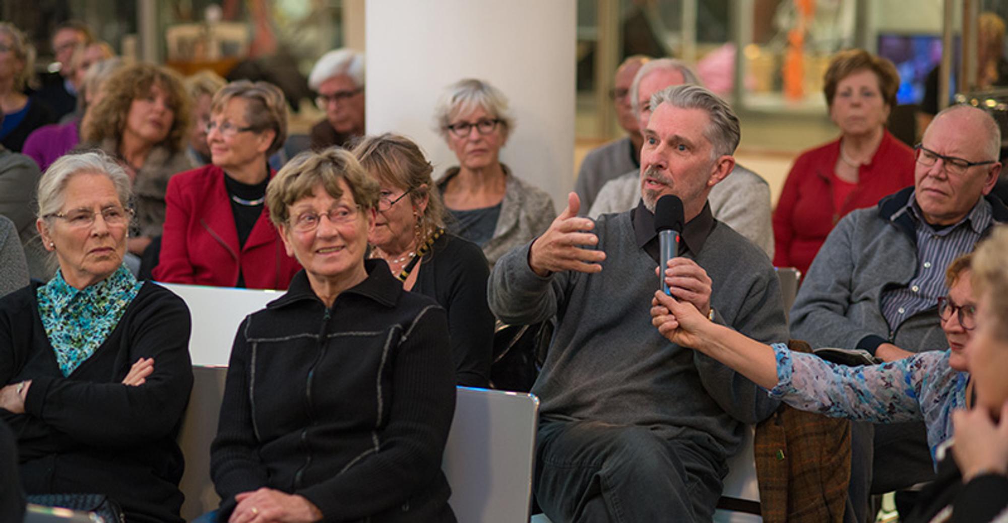 SCHUNCK debat - lezing Bezoekers zitten op stoelen in de centrale hal van Schunck. Een bezoeker uit het publiek praat door een microfoon.