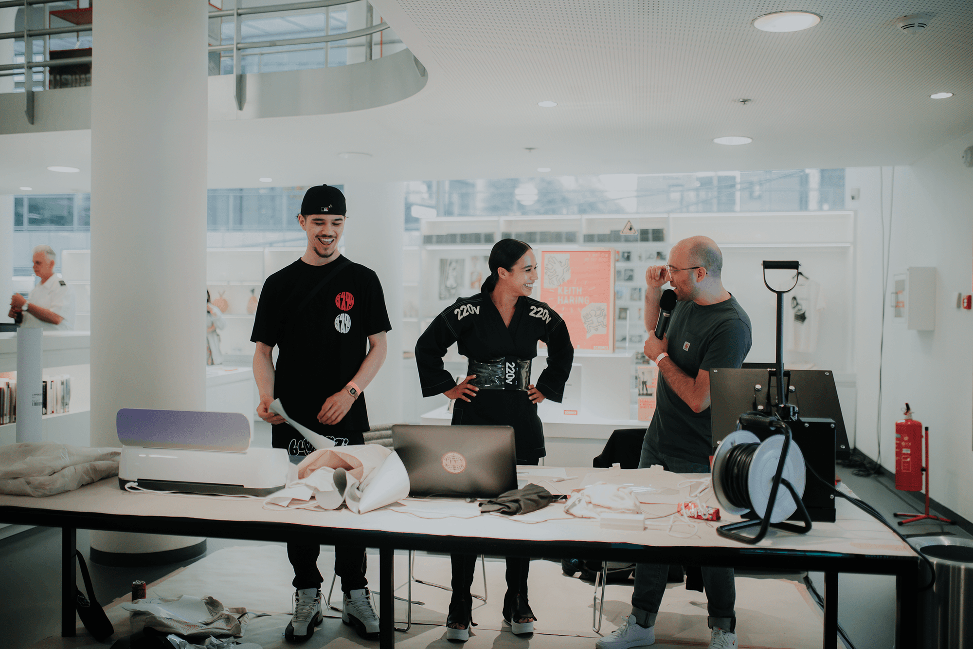 Drie mensen staan lachend bij een tafel vol materialen en laptops in een moderne ruimte; op de achtergrond posters over Keith Haring en een t-shirtdisplay.