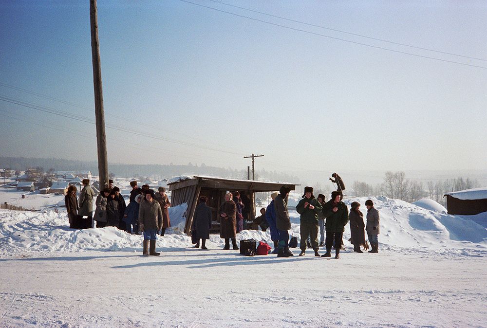 Fotografie 'Bus Stop' van Bertien van Manen (1994), Apanas, Siberië. Een groep mensen in winterkleding staat bij de bushalte in de sneeuw, met een blauwe lucht op de achtergrond.