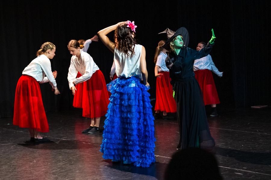 Groep mensen op een toneel, met centraal twee hoofdrolspelers: één in een blauwe gerafelde rok, witte top en roze bloem in het haar, en de ander in zwart met groene schmink, mogelijk als heks. Op de achtergrond staan andere performers in witte tops en rode rokken, tegen een donkere gordijnachtergrond.