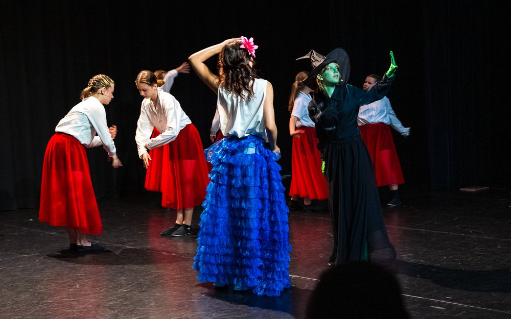 Groep mensen op een toneel, met centraal twee hoofdrolspelers: één in een blauwe gerafelde rok, witte top en roze bloem in het haar, en de ander in zwart met groene schmink, mogelijk als heks. Op de achtergrond staan andere performers in witte tops en rode rokken, tegen een donkere gordijnachtergrond.