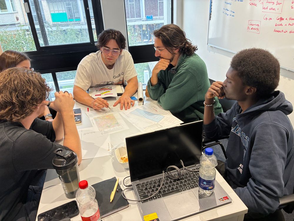 Vijf studenten zitten rond een tafel en bespreken gezamenlijk een architectuurtekening. Een deelnemer wijst met een potlood details aan op het ontwerp.
