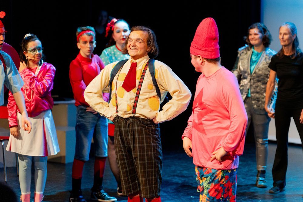 Groep performers staat op een podium in kleurrijke kostuums; twee spelers staan vooraan, één met bretels en één met een puntmuts, terwijl anderen op de achtergrond toekijken.