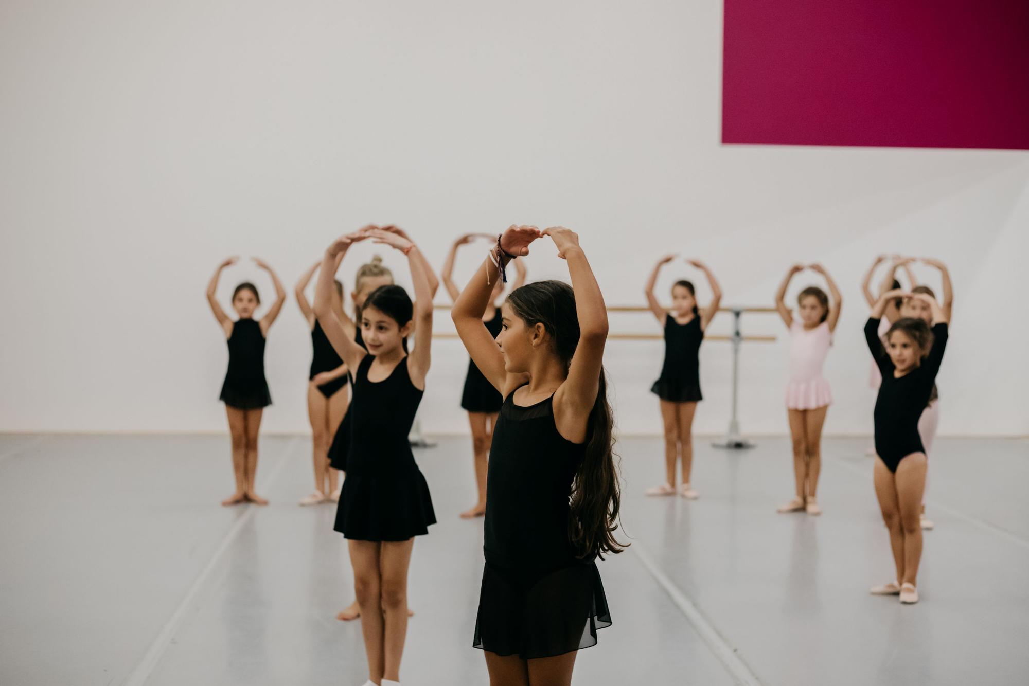 Groep jonge dansleerlingen oefent een balletpositie in een lichte dansstudio. Op de voorgrond staan twee leerlingen met de armen boven het hoofd, terwijl de rest van de groep dezelfde houding aanneemt.