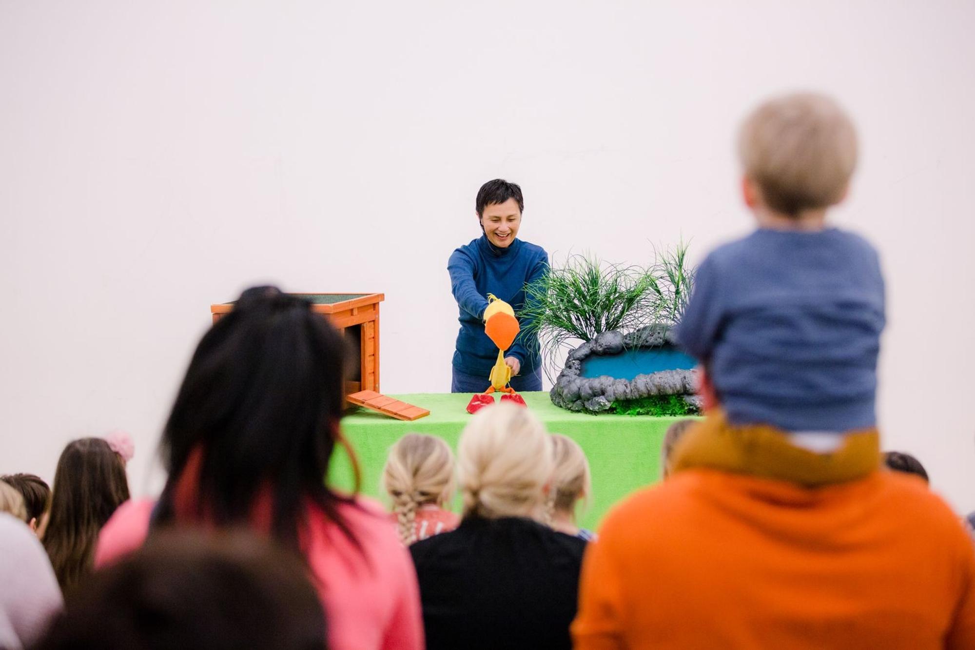 Artiest tijdens een peutervoorstelling met een eendenpop, terwijl kinderen en ouders kijken.