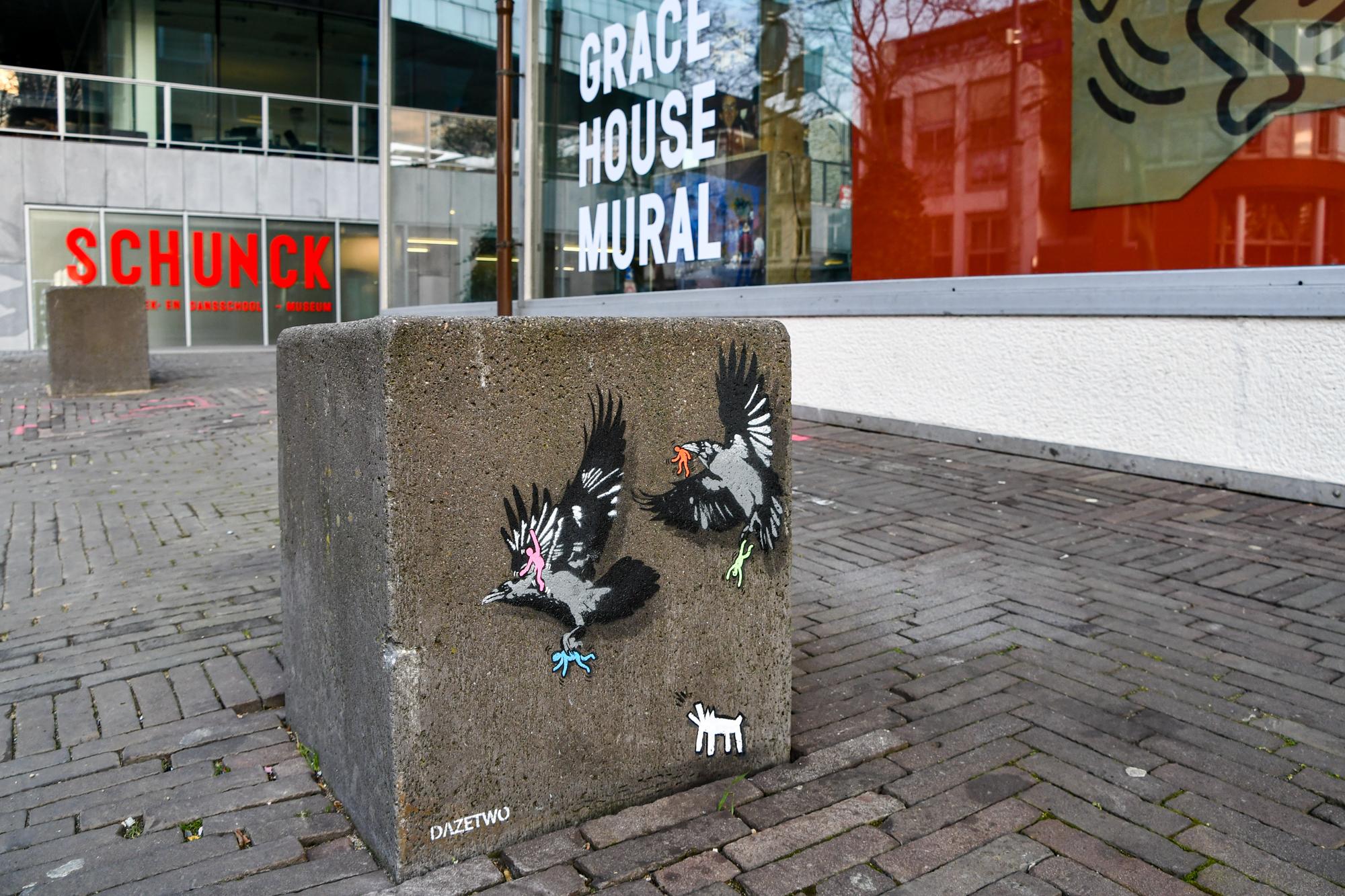 Een straatbeeld met een betonnen blok met graffiti van twee vogels en een klein wit dier. Op de achtergrond zijn gebouwen met borden waarop 'SCHUNCK' en 'GRACE HOUSE MURAL' staan.