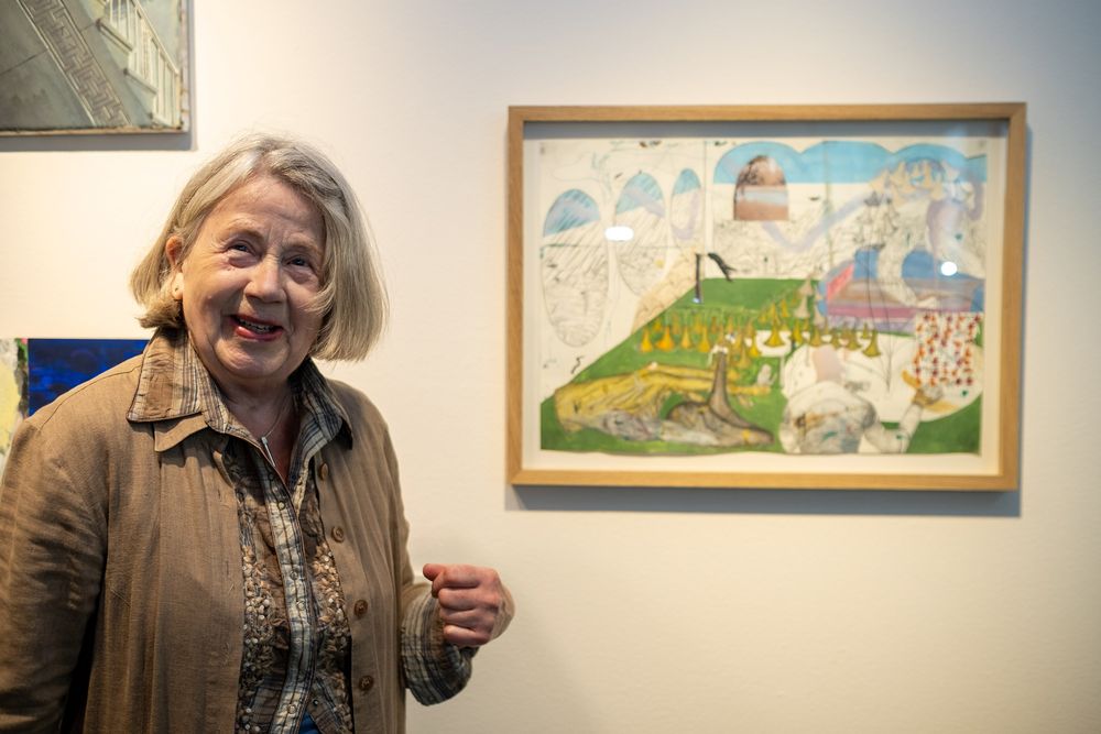 Een vrouw kijkt naar de camera, met links achter haar een kleurrijk schilderij dat de achtergrond siert.