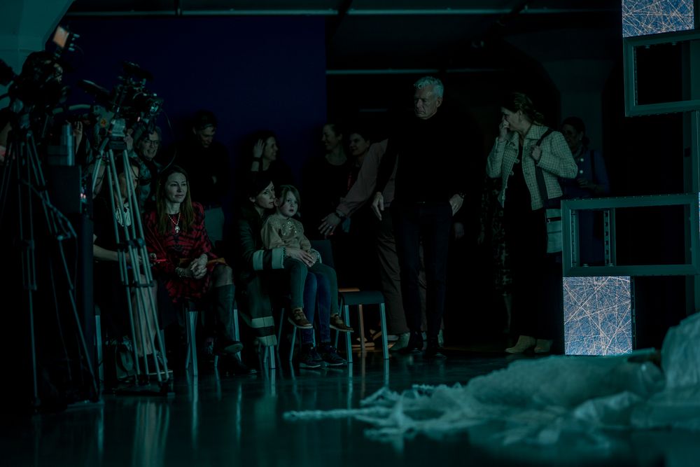 Bezoekers zitten en staan in een donkere ruimte met blauwe verlichting en camerastatieven, terwijl ze aandachtig naar een kunstpresentatie of optreden kijken; een kind zit op schoot bij een volwassene.