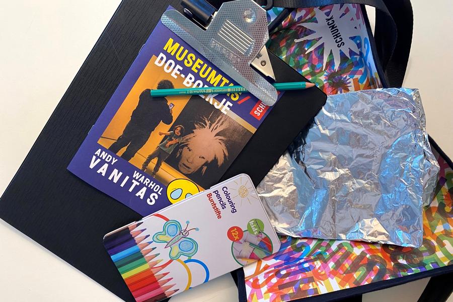 Een creatieve verzameling knutselmaterialen op tafel, waaronder het “Museumtas Doe-boekje” met Andy Warhol op de cover, kleurpotloden, aluminiumfolie, potloden, een clipboard en een kleurrijke tas met het SCHUNCK-logo.

