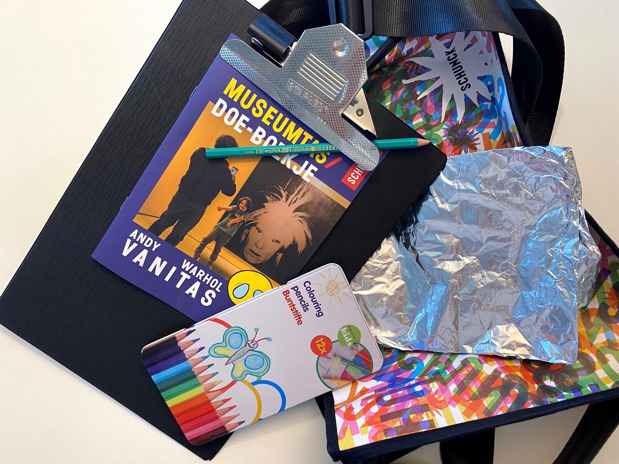 Een creatieve verzameling knutselmaterialen op tafel, waaronder het “Museumtas Doe-boekje” met Andy Warhol op de cover, kleurpotloden, aluminiumfolie, potloden, een clipboard en een kleurrijke tas met het SCHUNCK-logo.
