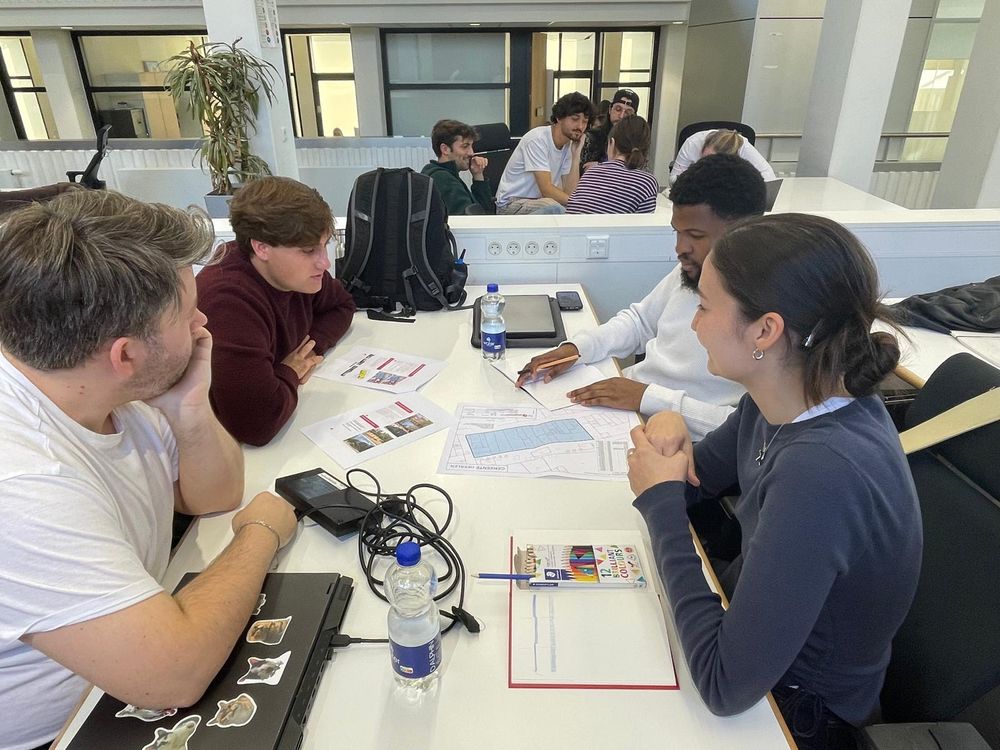 Vier studenten bespreken architectuurplannen aan een witte tafel. Op de achtergrond werken andere deelnemers in groepjes aan hun ontwerpen.