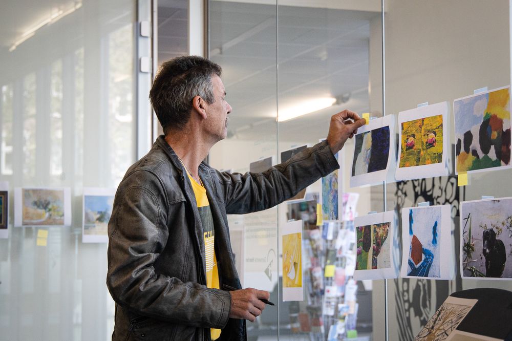 Een man hangt met een stukje tape een kunstwerk op aan een glazen wand, omringd door andere kleurrijke afbeeldingen.
