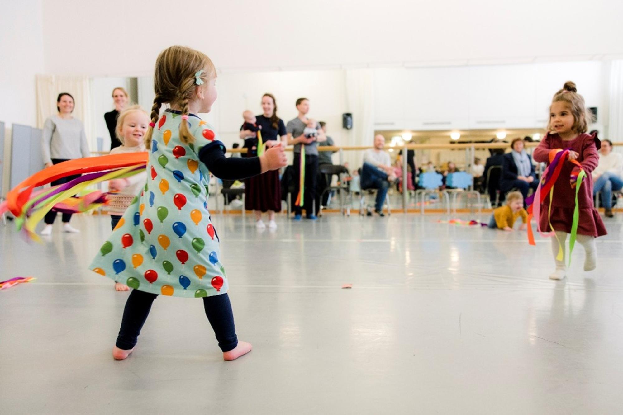 Twee jonge kinderen dansen vrolijk met gekleurde linten in een lichte dansstudio, terwijl ouders en begeleiders vanaf de achtergrond toekijken. De kinderen bewegen vrij op blote voeten en genieten zichtbaar van het spel.