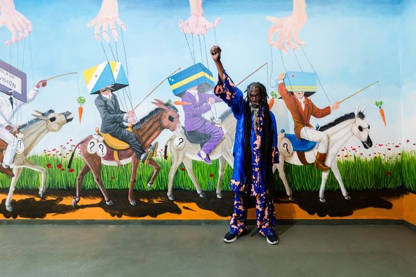 Kunstschilder Elijah Nicholas voor zijn muurschildering waarin marionetten en rijders op ezels kritiek leveren op koloniale machtsstructuren.