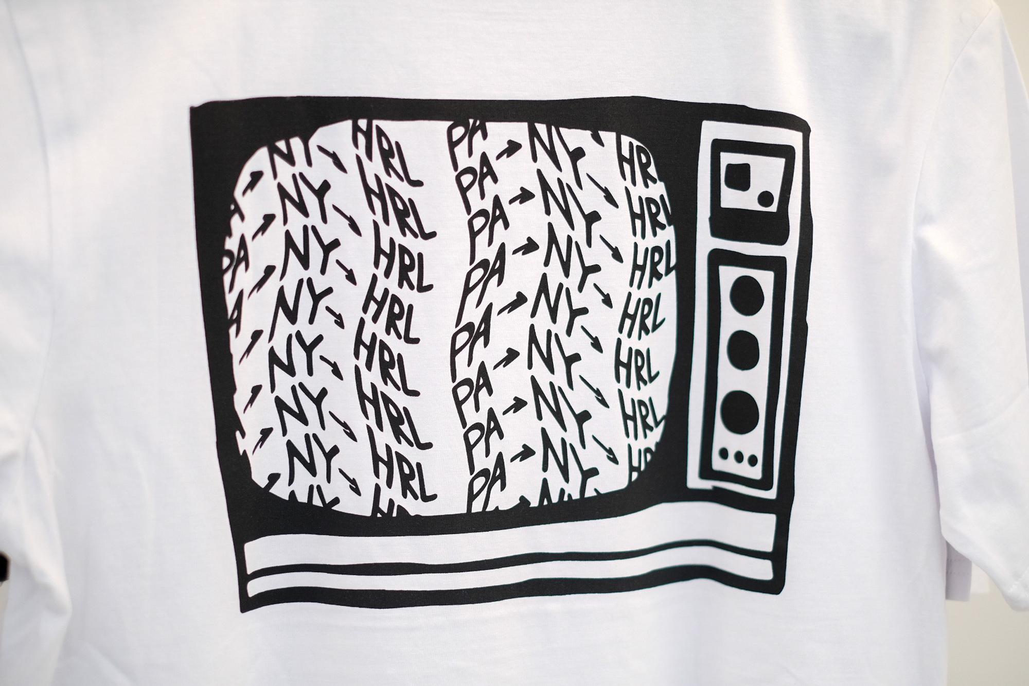 Wit t-shirt met een zwart grafisch ontwerp van een ouderwetse televisie. In het scherm van de televisie staan herhaalde tekstpatronen met 'NY,' 'PA,' en 'HRL.' De tekst is verspreid binnen het scherm. Het ontwerp bevat knoppen en draaiknoppen aan de rechterkant van de televisie.