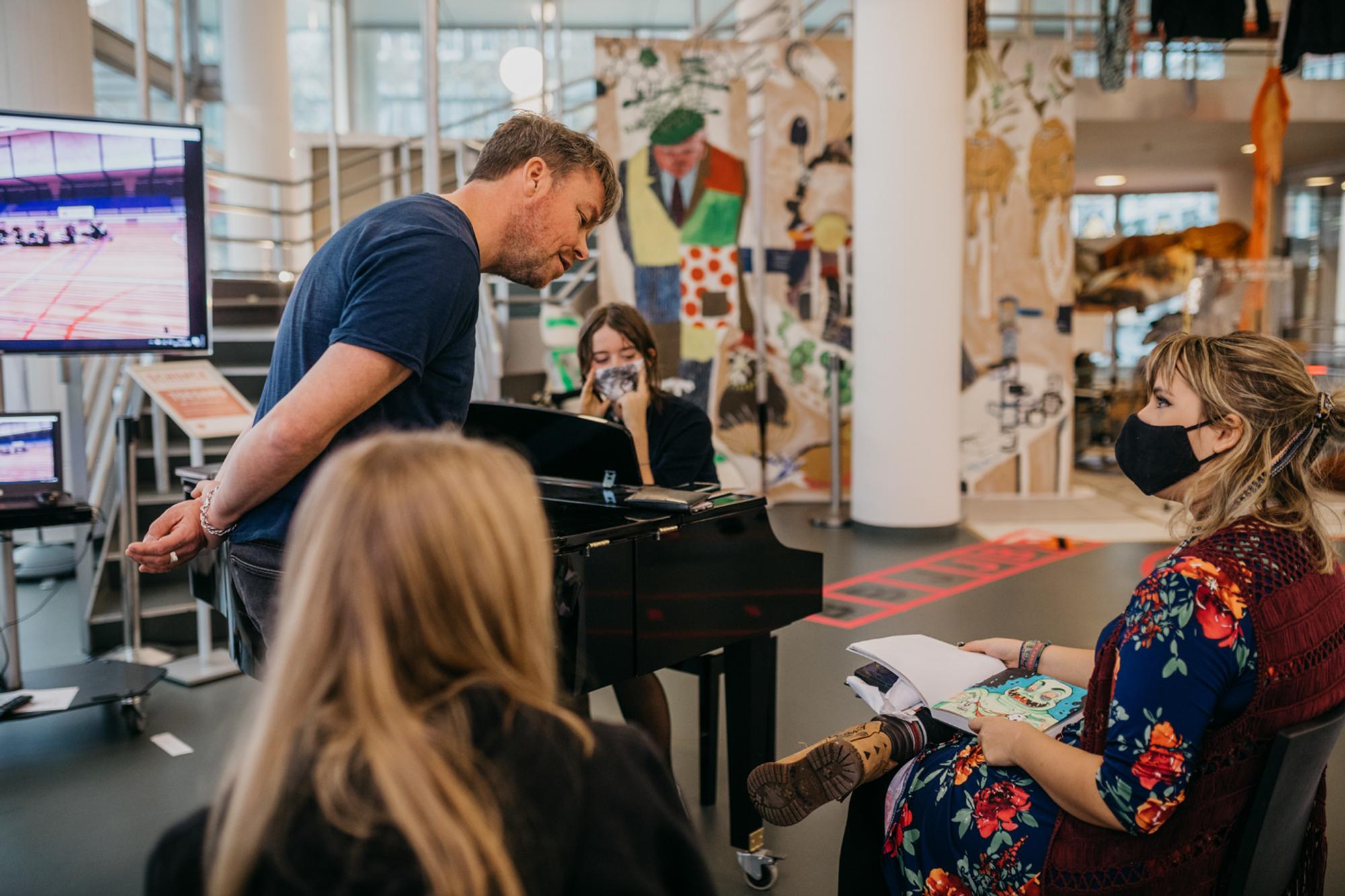 David Bade met drie vrouwen in de centrale hal van SCHUNCK. Op de achtergrond staat een scherm waarop een gymzaal te zien is en hangt een grote tekening. Eén van de vrouwen zit op een piano, ze dragen mondkapjes.
