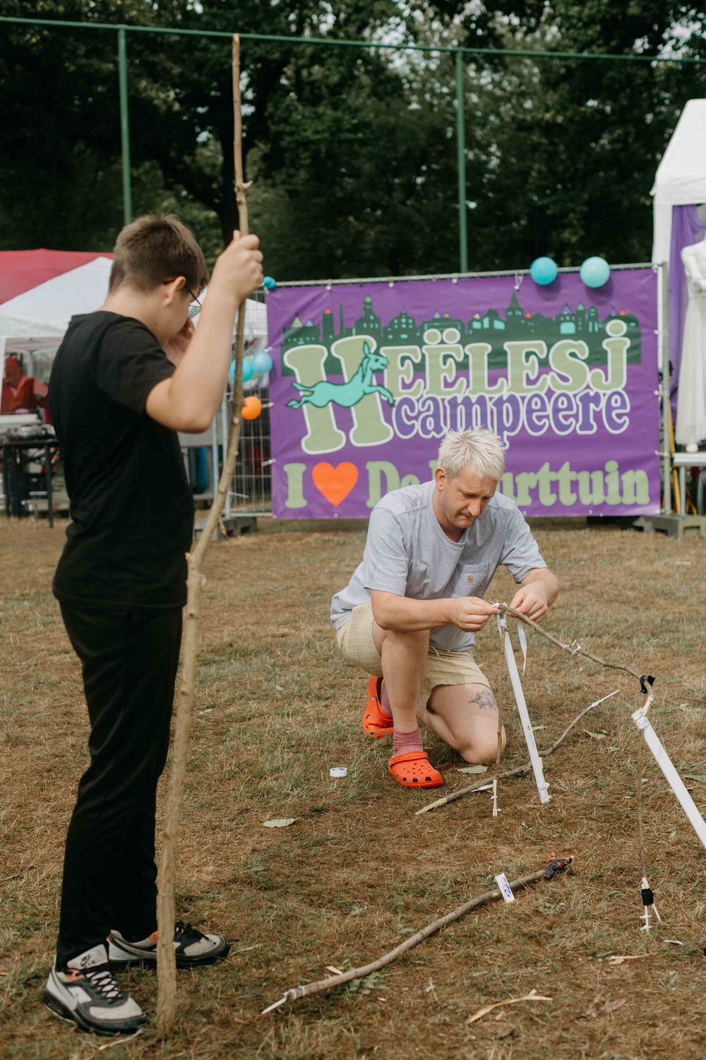Volwassene en kind versieren takken op een grasveld, met op de achtergrond een spandoek waarop "Heëlesj Campeere" staat.