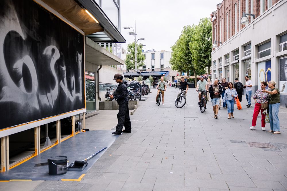 Straatscène met kunstenaar Martinus Papilaja die een muurschildering maakt, voorbijgangers kijken toe.