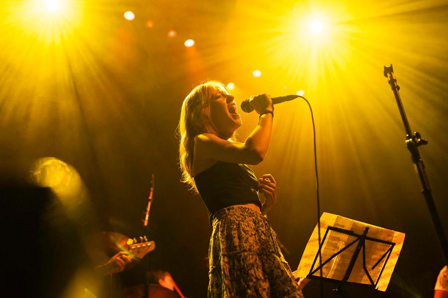 Een zangeres staat op een fel verlicht podium en zingt gepassioneerd in een microfoon, omringd door warm geel podiumlicht. Op de achtergrond zijn andere muzikanten en een muziekstandaard zichtbaar.