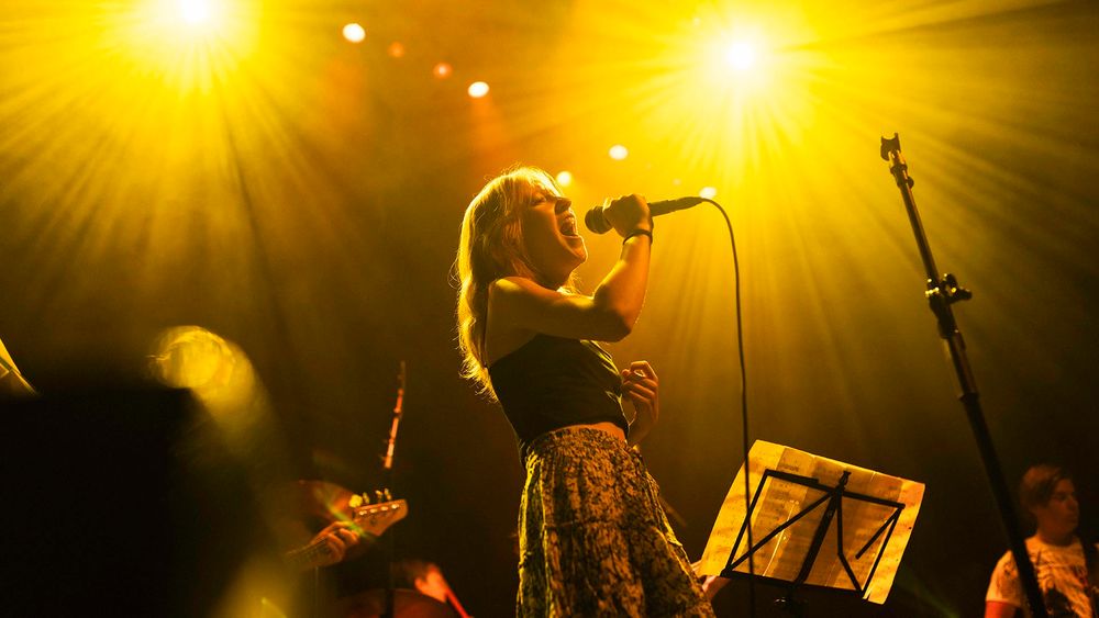 Een zangeres staat op een fel verlicht podium en zingt gepassioneerd in een microfoon, omringd door warm geel podiumlicht. Op de achtergrond zijn andere muzikanten en een muziekstandaard zichtbaar.