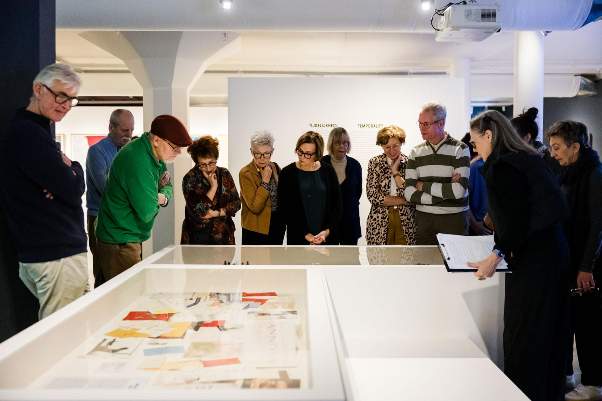 Groep mensen luistert aandachtig naar een rondleider in een museum, rondom een vitrinekast met documenten.