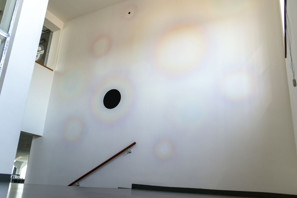 Een interieur met een witte muur waarop verschillende cirkelvormige, regenboogkleurige lichtreflecties te zien zijn. Er is een zwart cirkelvormig object op de muur en een houten leuning die naar beneden loopt. De vloer is grijs en er is een gedeeltelijk zicht op een andere kamer of ruimte door een opening aan de linkerkant van de afbeelding.