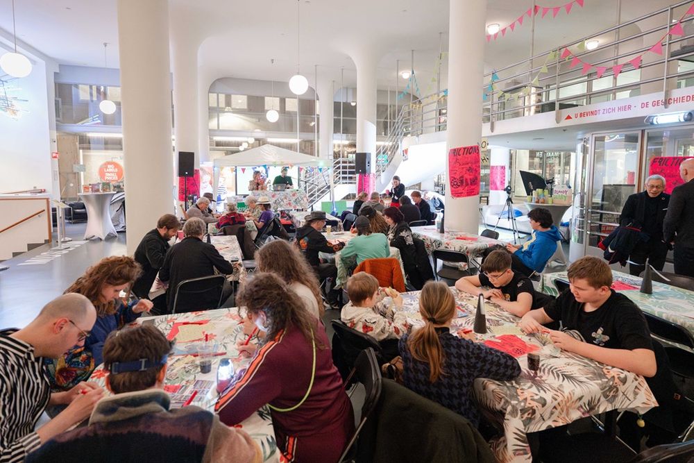 Deelnemers van verschillende leeftijden zitten aan lange tafels in een lichte ruimte met witte zuilen en hoge plafonds, verdiept in een gezamenlijke knutsel‑ of kunstactiviteit tijdens Frictions Festival #12; de tafels zijn bedekt met fleurige doeken, en op de achtergrond hangen roze vlaggetjes en posters terwijl mensen geconcentreerd werken en praten.