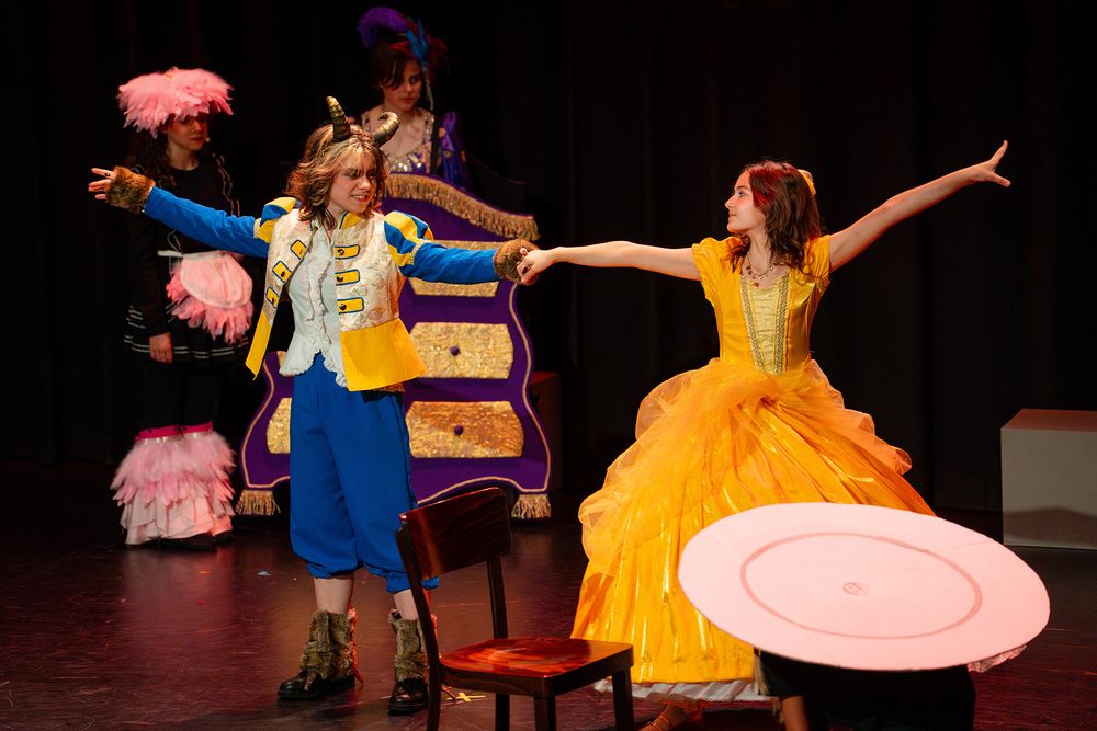 Twee performers staan op een podium tijdens de musical Belle en het Beest terwijl ze elkaars hand vasthouden en met gestrekte armen naar buiten draaien. Eén performer draagt een felgele baljurk en de ander een blauw kostuum met een jas met gouden details, terwijl op de achtergrond decorstukken, een stoel en andere kostuums zichtbaar zijn.
