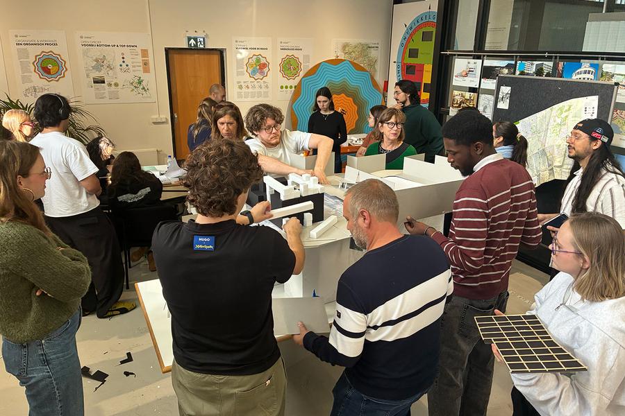 Een groep deelnemers bekijkt en bespreekt een grote architectuurmaquette met witte en zwarte gebouwen. Op de achtergrond hangen kaarten, tekeningen en presentaties.