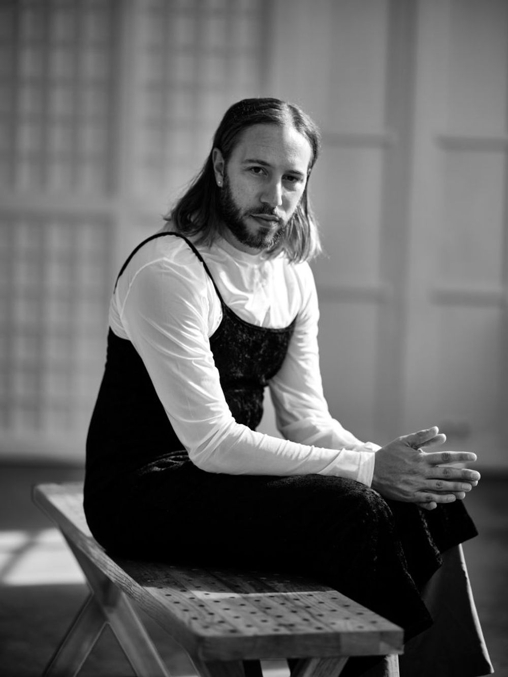Zwart-wit portret van een persoon met halflang haar en baard, zittend op een bankje. De persoon draagt een licht shirt met daarover een donker kledingstuk en kijkt recht in de camera.