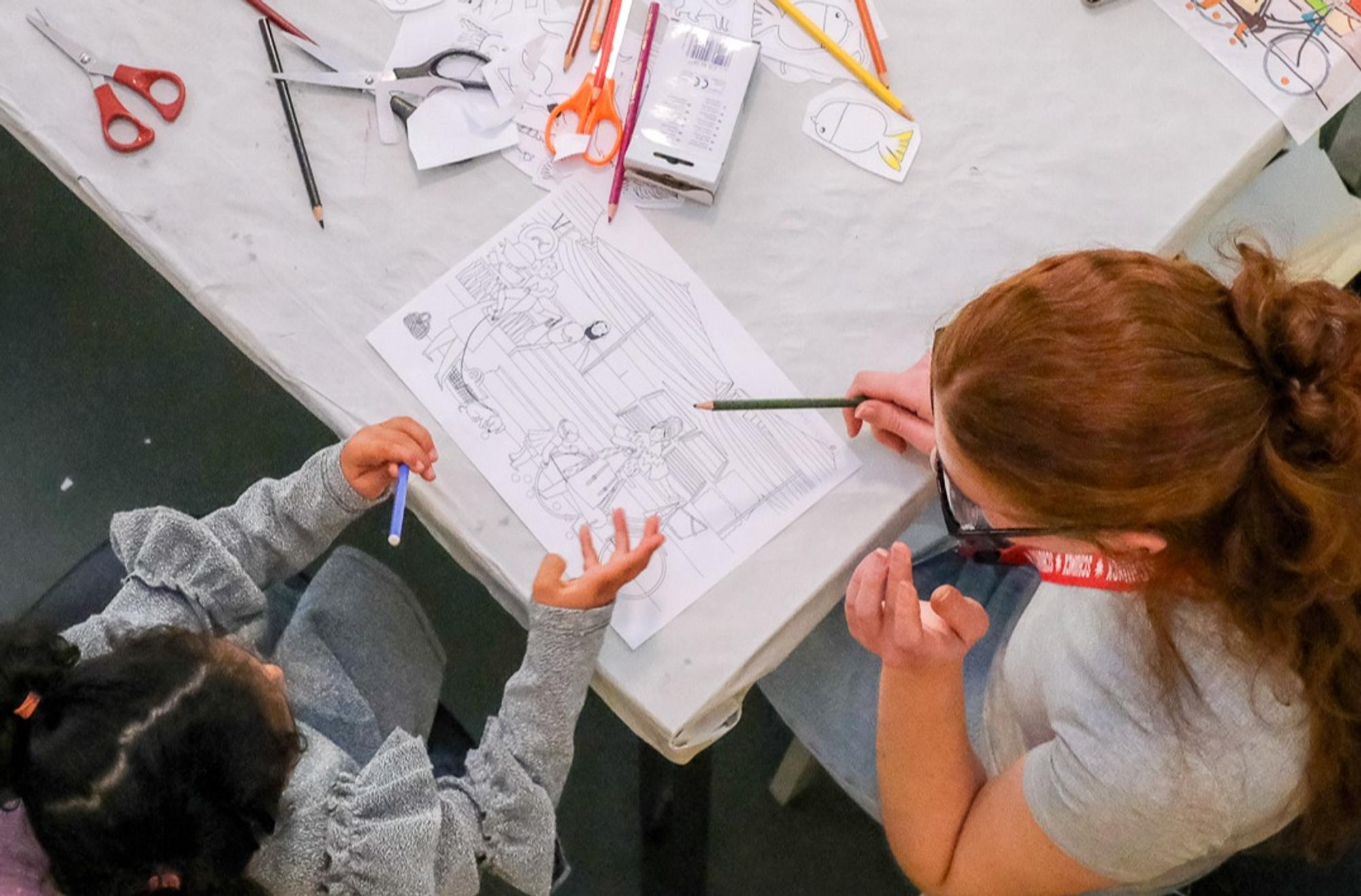 Tijdens een activiteit rond vrijheid en verhalen maken een begeleider en een kind samen een tekening aan een tafel vol kleurpotloden, scharen en knutselmateriaal. Vanuit vogelperspectief is te zien hoe deelnemers creatief aan de slag gaan met een opdracht die aansluit bij het programma van het Vrijheidsdiner.
