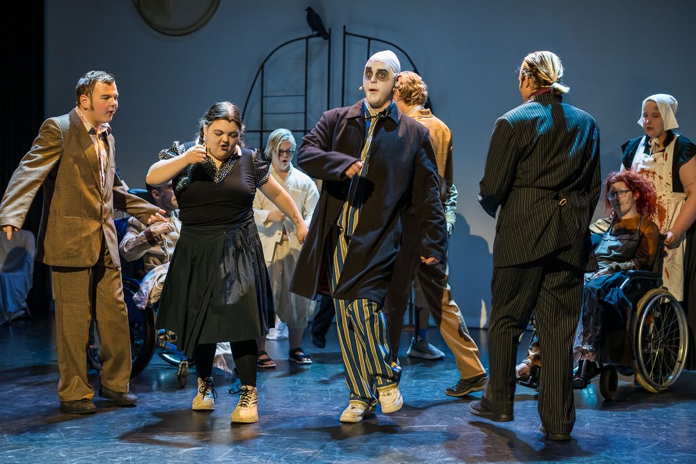 Groep mensen op een toneel tijdens een theatervoorstelling, gekleed in diverse kostuums. Eén persoon zit in een rolstoel. Op de achtergrond zijn toneelattributen en verlichting zichtbaar.