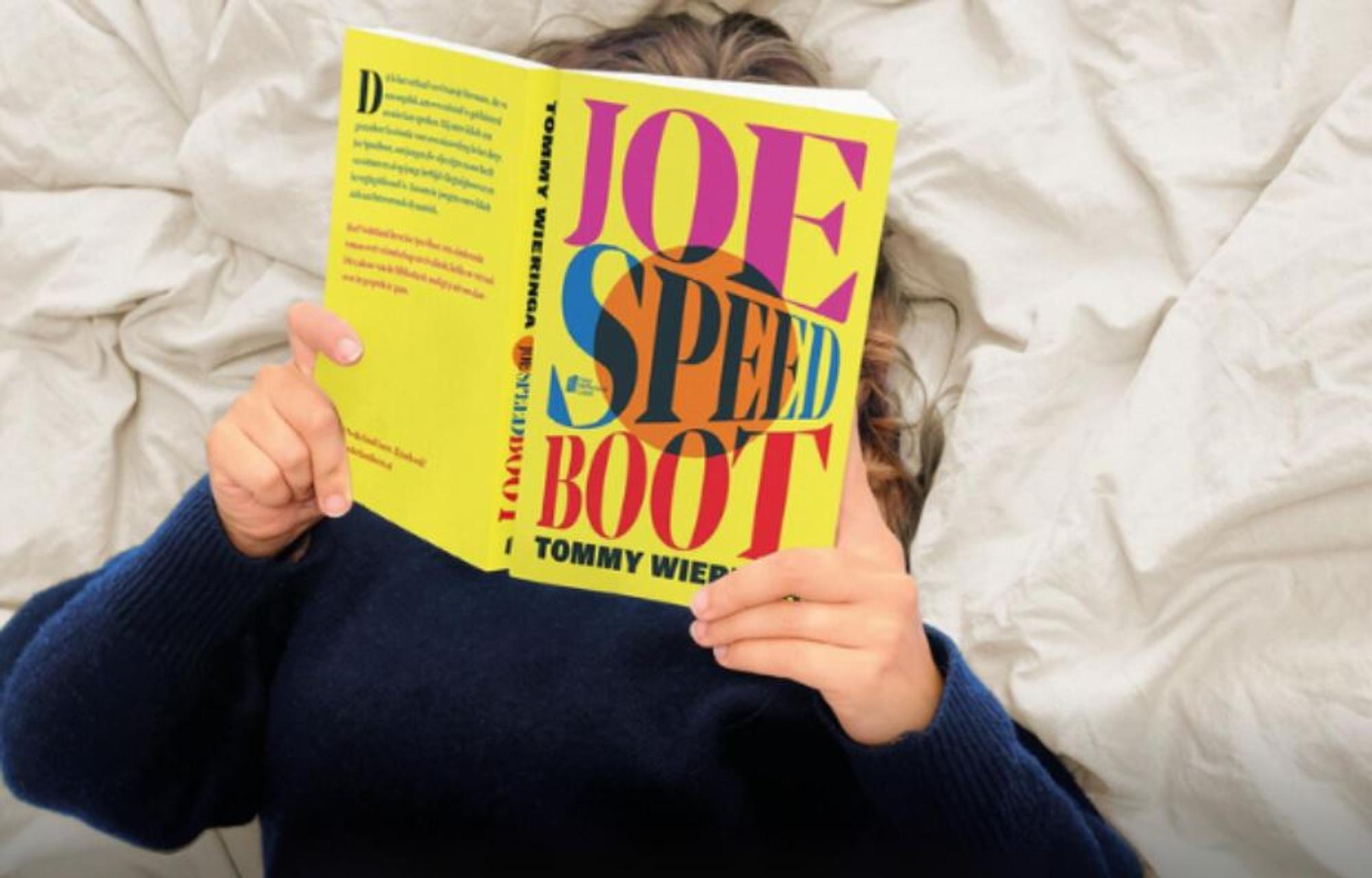 Iemand die op bed ligt op zijn rug met een boek boven zijn hoofd genaamd 'Joe speed boot' .
