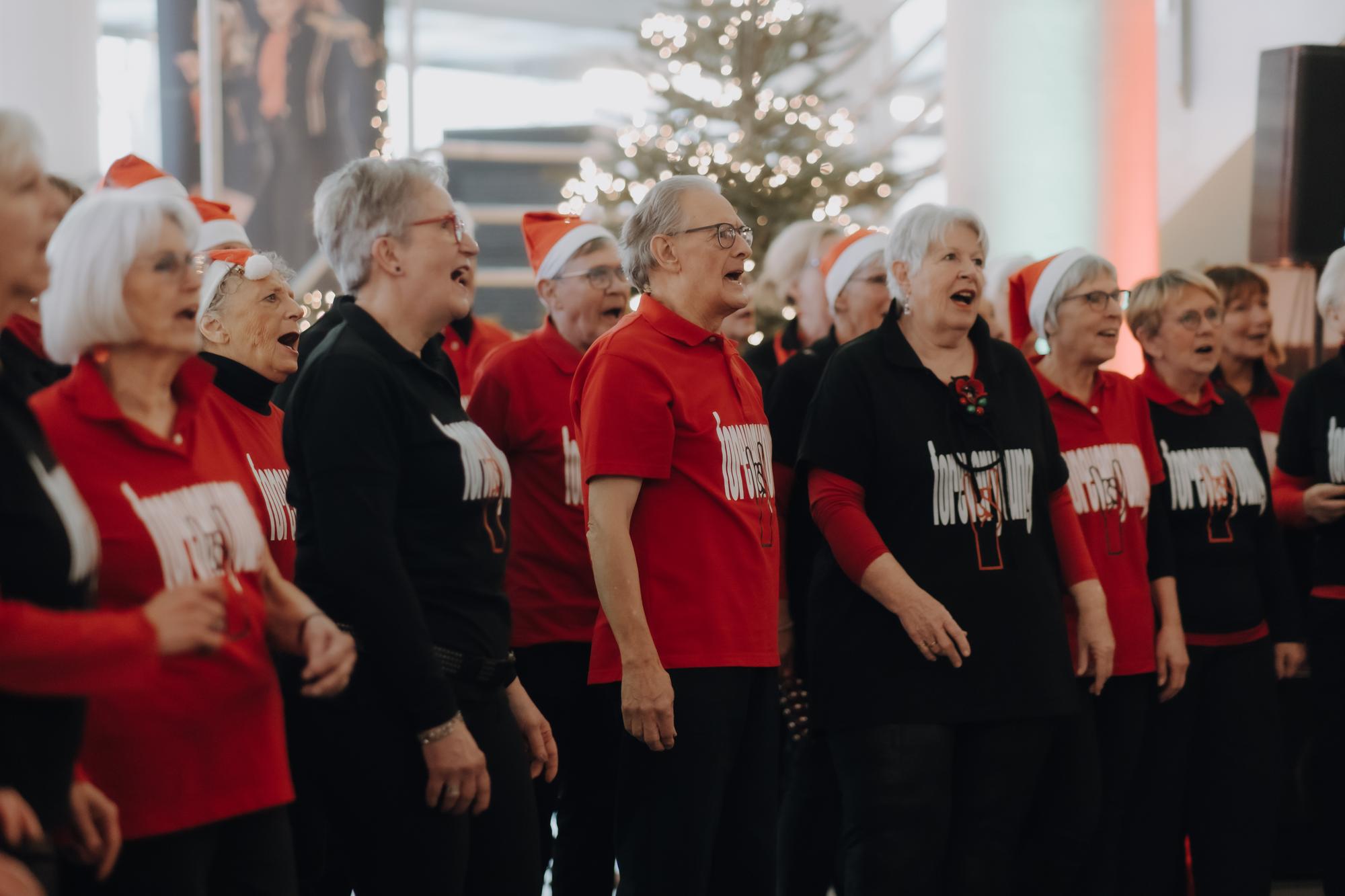Mannen en vrouwen uit een koor, gekleed in zwart en rood, zingen en treden op in de hal van SCHUNCK.