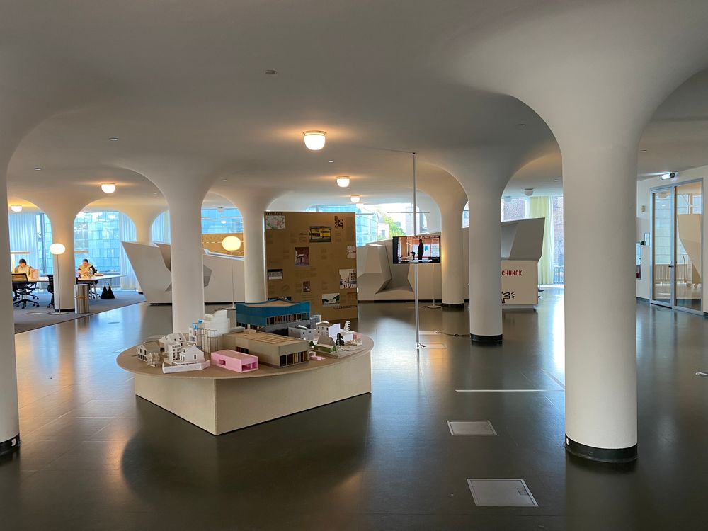 Interieur van het Glaspaleis in Heerlen met witte pilaren en een open, lichte ruimte. In het midden staat een ronde tafel met architectuurmaquettes van gebouwen, terwijl op de achtergrond panelen met informatie en bezoekers te zien zijn.